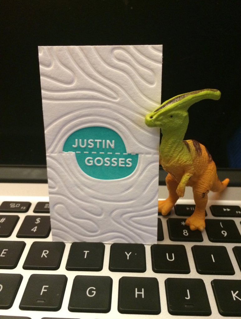 "A photograph of a toy dinosaur holding Justin Gosses's business card."